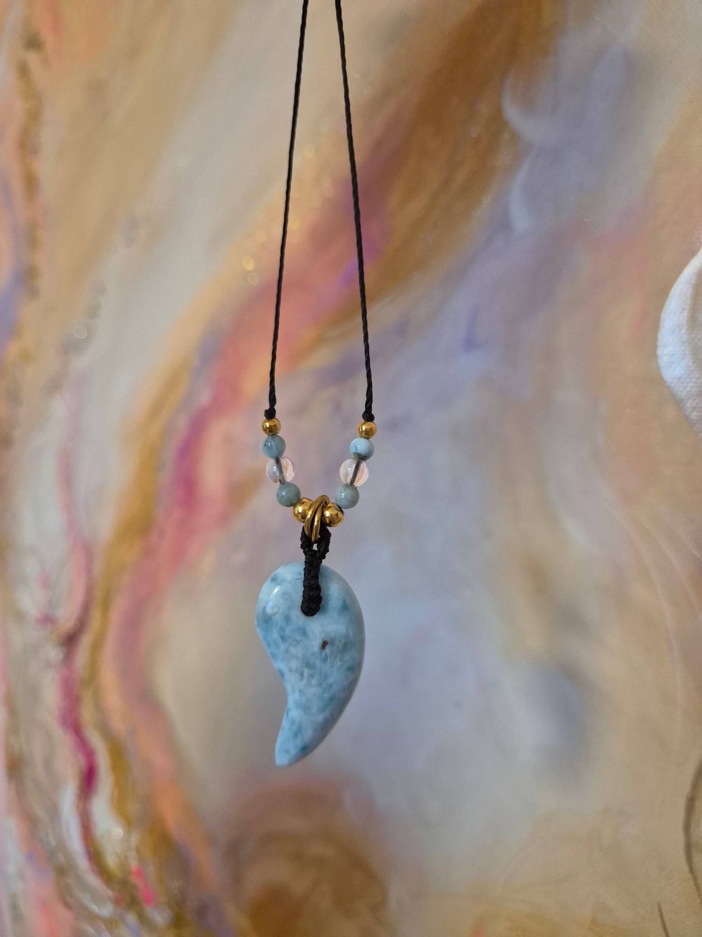 Handgefertigter Magatama-Anhänger aus hellblauem Larimar-Edelstein an einer Makramee-Kette, dekoriert mit kleinen Perlen und als Diffuser für ätherische Öle oder Parfum. Das japanische Symbol für Lebenskraft und Schutz liegt auf einem naturfarbenen Stoffhintergrund neben einem Lavastein.