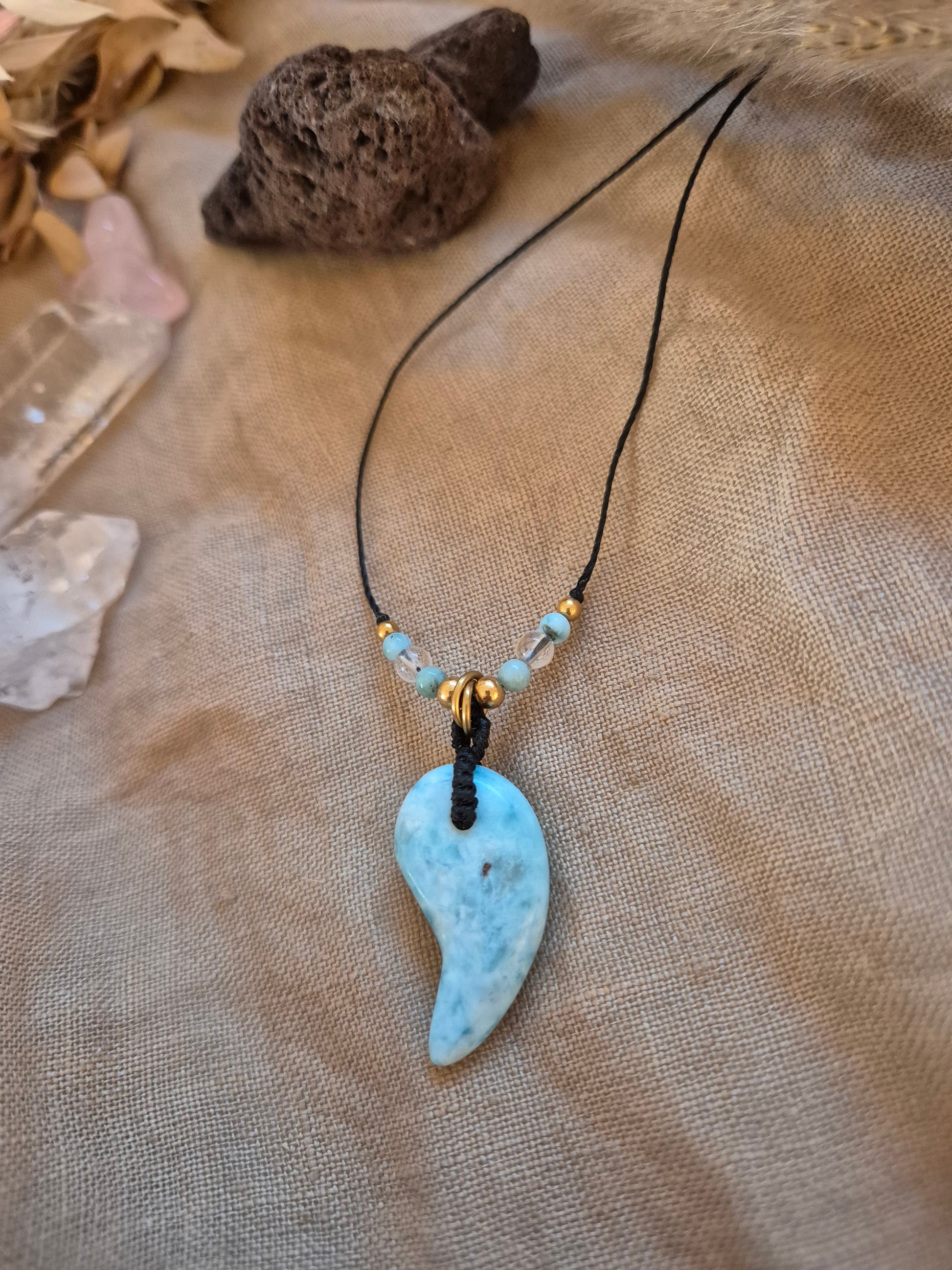 Handgefertigter Magatama-Anhänger aus hellblauem Larimar-Edelstein an einer Makramee-Kette, dekoriert mit kleinen Perlen und als Diffuser für ätherische Öle oder Parfum. Das japanische Symbol für Lebenskraft und Schutz liegt auf einem naturfarbenen Stoffhintergrund neben einem Lavastein. 