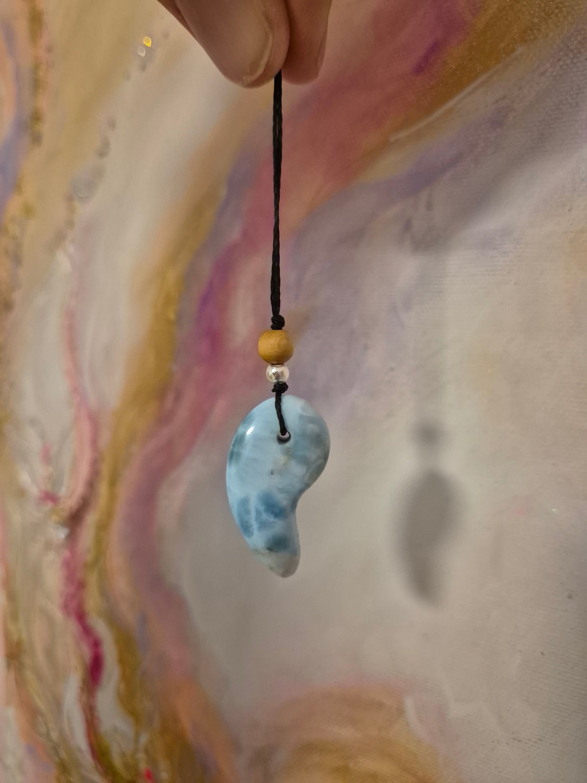 Handgefertigter Magatama-Anhänger aus hellblauem Larimar-Edelstein an einer Makramee-Kette, dekoriert mit kleinen Perlen und als Diffuser für ätherische Öle oder Parfum. Das japanische Symbol für Lebenskraft und Schutz liegt auf einem naturfarbenen Stoffhintergrund neben einem Lavastein. 