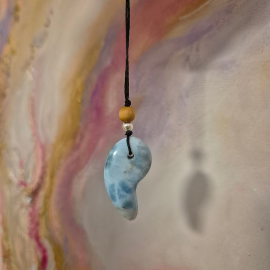 Handgefertigter Magatama-Anhänger aus hellblauem Larimar-Edelstein an einer Makramee-Kette, dekoriert mit kleinen Perlen und als Diffuser für ätherische Öle oder Parfum. Das japanische Symbol für Lebenskraft und Schutz liegt auf einem naturfarbenen Stoffhintergrund neben einem Lavastein. 